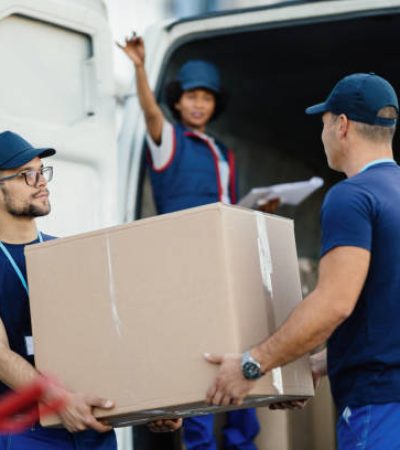 Delivery men cooperating while loading cardboard boxes in a mini van. Their female colleagues is gesturing from a van.